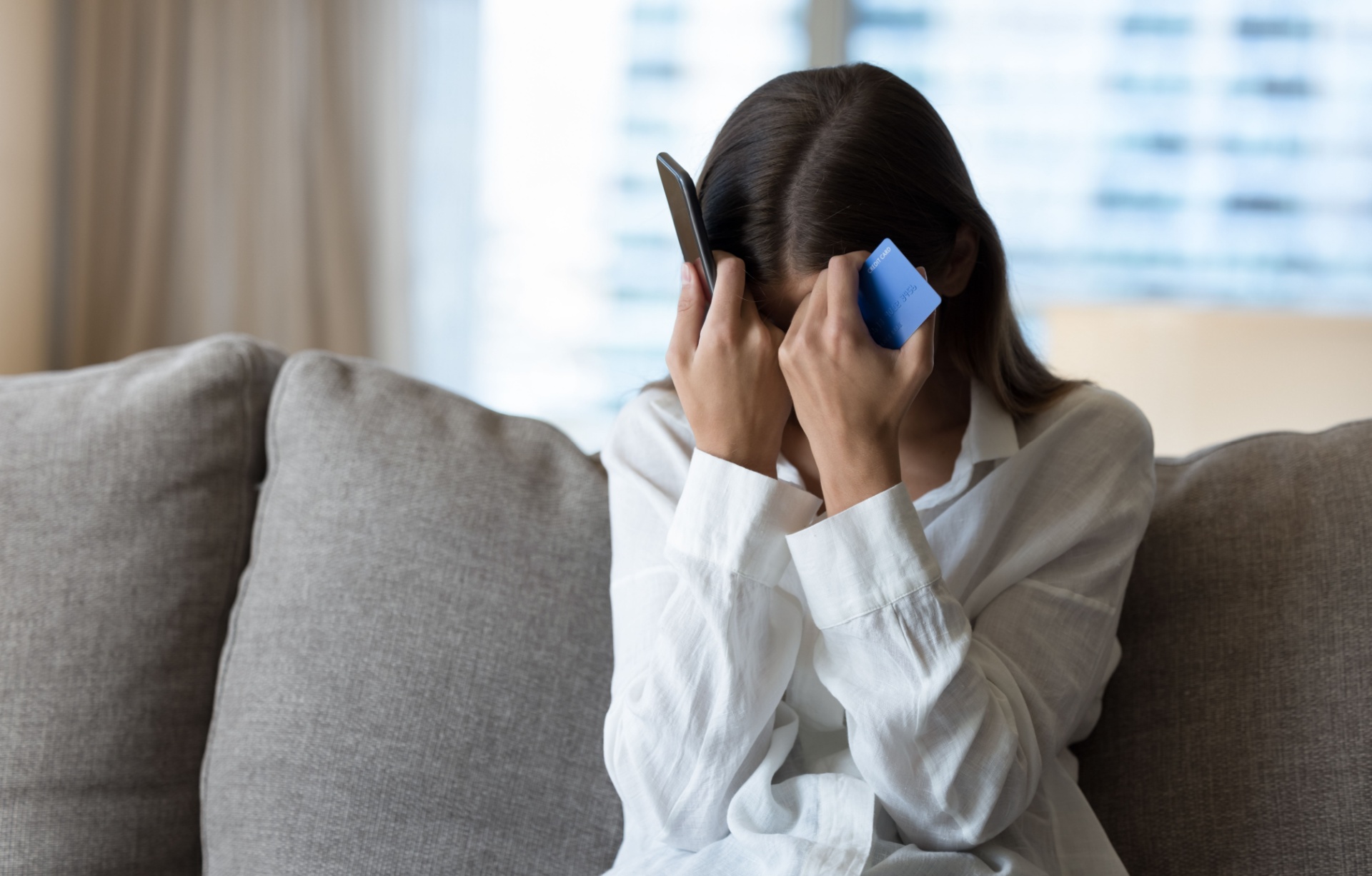 A woman holds her head with a phone in one hand and a bank card in the other.